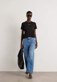 Svart bomull t-skjorte med et lite logo, lyseblå rette jeans, svart håndveske, og svarte åpen-tå sandaler. Modellen står mot en nøytral bakgrunn.