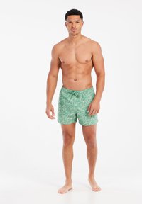 Gröna badshorts med ett vitt bladmönster, elastisk midja med dragsko och längd ovanför knäna. Inga synliga fickor.