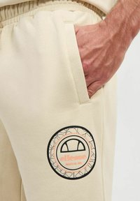Pantalones de chándal beige con un acabado texturizado, que presentan un logotipo gráfico redondo en negro y rosa en el muslo y un bolsillo lateral.