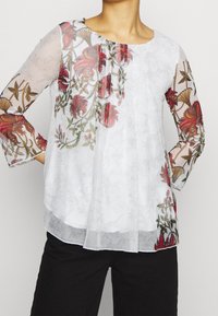 Blusa blanca de tela transparente con bordado floral rojo en las mangas y el frontal. Escote redondeado, silueta fluida.