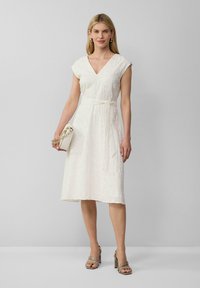 Robe blanche avec un décolleté en V, de courtes manches à capuchon, et une ceinture à la taille. Ourlet surélevé et motif floral texturé. Associée à des talons neutres.
