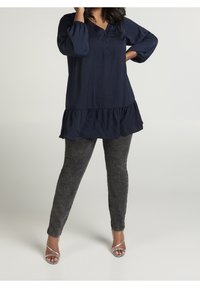 Mörkblå tunika med V-ringning, tre knappar och volangslut. Kombinerad med strukturerade svarta slim fit-byxor och silverfärgade klacksandaler.