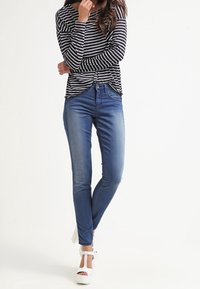 Pruhovaný dlouhý rukáv top spárovaný s přiléhavými modrými džíny a bílými sandály na platformě. Tkanina vypadá lehce a má hladkou texturu.