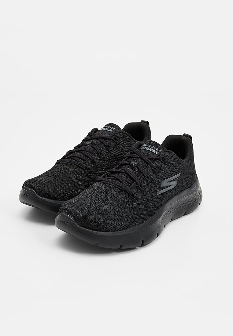 Zapatillas deportivas de malla negra con puntera redondeada, parte superior texturizada y suela acolchada. Presentan un diseño con cordones y el logo de Skechers en el costado.