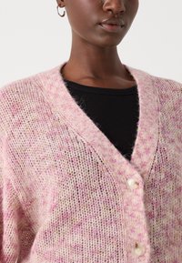 Cardigan lavorato a maglia di un morbido rosa con leggere sfumature beige. Presenta un profondo scollo a V, una vestibilità oversized e due bottoni sulla parte anteriore.