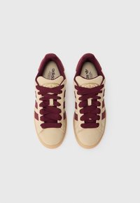 Bēša un bordō krāsas Adidas sporta kurpes ar bordō krāsas šņorēm, skatītas no augšas uz baltas fona.