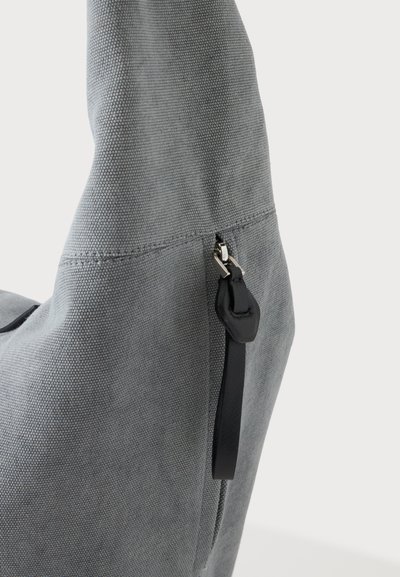 Gros plan d’un sac en tissu gris avec un curseur de fermeture éclair en cuir noir sur une fermeture éclair visible sur un fond clair uni.