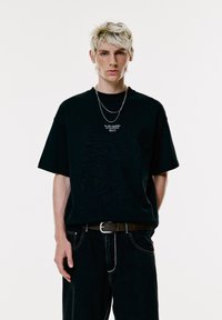Giovane con capelli biondi corti, che indossa una t-shirt nera oversize con testo bianco e catene argento sovrapposte, in piedi davanti a uno sfondo bianco semplice.