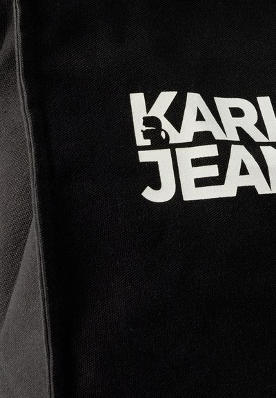 Черен плат с дебели бели букви „KARL JEAN“ и малък силует на човек с очила, вплетен в буквата „K“.