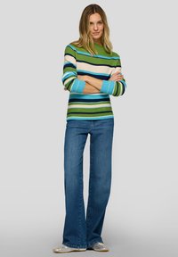 Grüner und blauer gestreifter Strickpullover mit gerippten Bündchen, kombiniert mit dunkelblauen Jeans mit geradem Bein. Das Model steht mit verschränkten Armen.