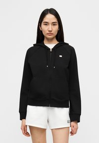 Μαύρο φούτερ με φερμουάρ από μαλακό ύφασμα, με κουκούλα, μπροστινές τσέπες και ένα μικρό λογότυπο. Συνδυασμένο με λευκά αθλητικά σορτς.