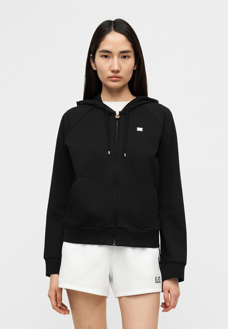 Μαύρο φού τερ με φερμουάρ από μαλακό ύφασμα, με κουκούλα, μπροστινές τσέπες και ένα μικρό λογότυπο. Συνδυασμένο με λευκά αθλητικά σορτς.