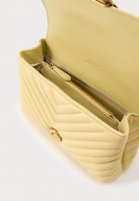 Κίτρινη δερμάτινη τσάντα με ραφές και κούμπωμα με χρυσό κουμπί, διαθέτει εσωτερικό φερμουάρ και λείο εσωτερικό ύφασμα.