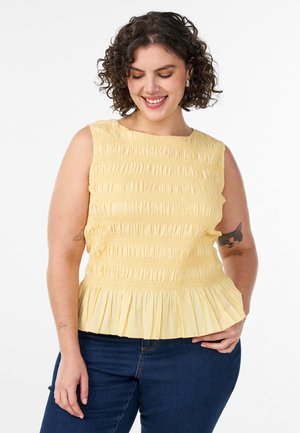 Kvinde med krøllet hår iført en gul ærmeløs top med rynkede detaljer og mørkeblå jeans, smilende og kiggende ned med hånden på hoften.