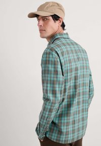 Turquoise plaid shirt with brown and yellow accents, featuring long sleeves and a buttoned cuff. Worn with a tan cap.