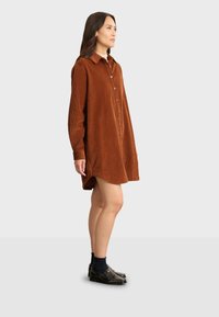 Braunes Cordhemdkleid mit durchgehender Knopfleiste, langen Ärmeln und abgerundetem Saum. Getragen mit schwarzen Loafers und dunklen Socken.