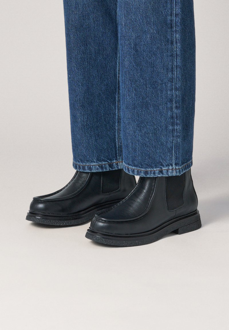 Botas de cuero negras hasta el tobillo con paneles elásticos en los lados, puntas redondeadas y suelas de goma texturizadas. Combinadas con pantalones de mezclilla azules.