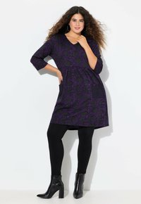 Robe violette avec motif floral noir, décolleté en V, manches 3/4, deux poches latérales, assortie à des leggings noirs et des bottines à talons.
