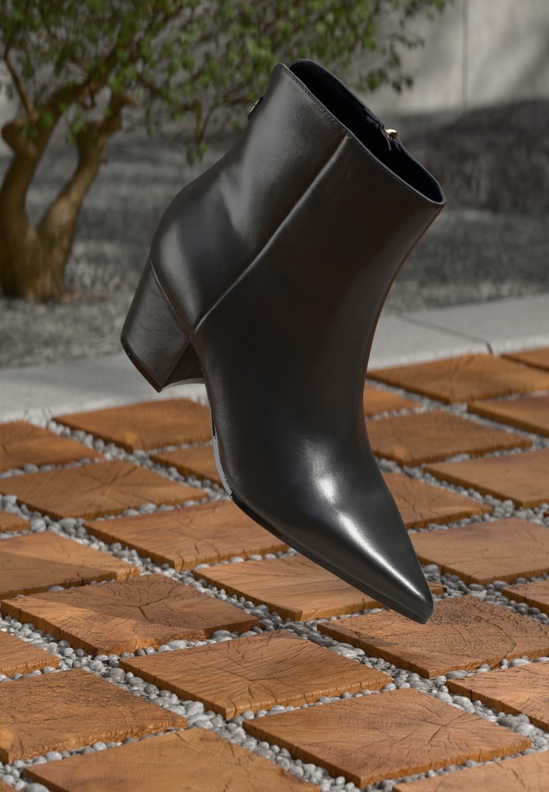 Bota de tornozelo em couro preto com bico apontado e salto moderado, apresentando uma textura suave e detalhes mínimos, em um fundo estampado.