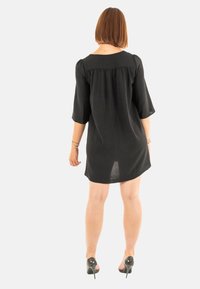 Vestido negro a la altura de la rodilla con mangas de tres cuartos, corte holgado y detalle de plisado en la parte posterior. Combinado con zapatos de tacón alto negros.