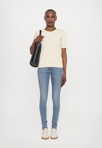 Camisa de manga corta color beige claro, jeans ajustados azules y zapatillas beige; sosteniendo un bolso cilíndrico negro con una correa.