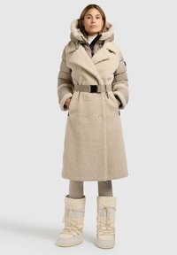 Beige dubbele rij knopen jas met een pluizige textuur, contrasterende gewatteerde mouwen, een oversized capuchon en een bijpassende riem. Witte sneeuwlaarzen maken de look compleet.