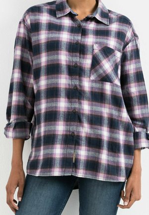 Femme portant une chemise à manches longues à carreaux bleu marine, rose et blanc, avec une poche poitrine et les manches retroussées, associée à un jean bleu.