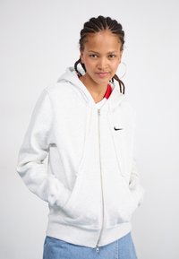 Lys grå zip-up hettegenser med frontlommer og snorhette. Har en liten svart Nike-logo på brystet. Materiale i bomullsblanding.