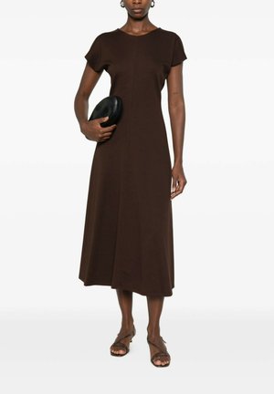 Donna che indossa un vestito midi marrone a maniche corte, tiene una pochette nera, abbinata a sandali marroni con cinturini su uno sfondo semplice.
