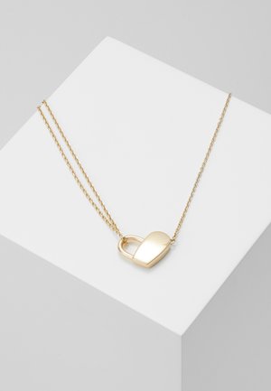Delikatny złoty naszyjnik z zawieszką w kształcie serca zamka, leżący na białej geometrycznej powierzchni.