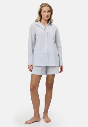 Kobieta stoi boso, ubrana w jasnoniebieską koszulę zapinaną na guziki z długim rękawem oraz pasujące szorty na jednolitym tle.