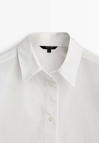 Chemise blanche à col boutonné avec deux boutons fermés, dotée d'un col pointu classique et d'une étiquette de marque visible à l'intérieur de l'encolure.