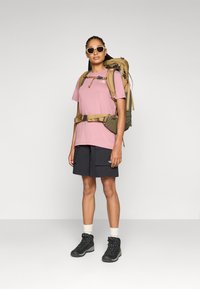 Rosa t-shirt med kort ärm, svarta cargo-shorts, svarta vandringskängor med snörning, stor grön ryggsäck med remmar och vita solglasögon.