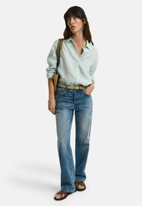 Lichtgroen overhemd met knopen, blauwe denim jeans, gevlochten beige riem, platte bruine sandalen en een tan schoudertas.