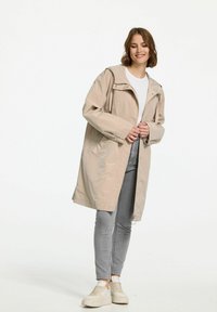 Beige waterdichte parka met capuchon, vooraan met ritssluiting, zijzakken en verstelbare trekkoorden; gedragen over een licht shirt en grijze jeans.