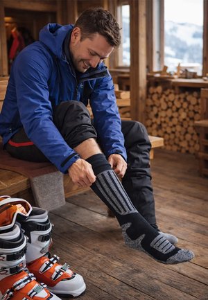 Mann in blauer Jacke sitzt drinnen auf einer Holzbank, zieht Skisocken an, daneben stehen Skischuhe auf dem Boden.