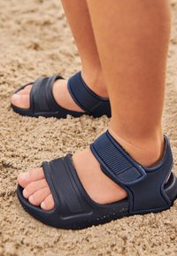 Sandali blu navy con spesse cinghie, realizzati in materiale flessibile, dotati di chiusure in velcro e una suola testurizzata, posizionati su terreno sabbioso.
