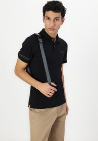 Čierne polo tričko s krátkym rukávom, na ktorých sú logá "GUESS" na manžetách a s úzkym strihom. Spárované s béžovými nohavicami a taškou s ramenným popruhom.