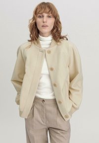 Beige Fleece-Jacke mit großen Knöpfen, rundem Kragen und lockerer Passform. Kombiniert mit einem weißen Rollkragenpullover und hellbraunen, maßgeschneiderten Hosen.