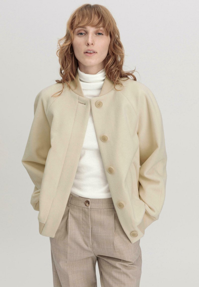 Beige Fleece-Jacke mit großen Knöpfen, rundem Kragen und lockerer Passform. Kombiniert mit einem weißen Rollkragenpullover und hellbraunen, maßgeschneiderten Hosen.
