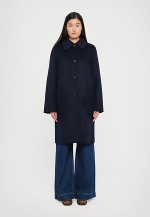 Cappotto di lana blu navy con colletto a punta, cinque bottoni e due tasche frontali, abbinato a pantaloni di jeans a gamba larga e scarpe marroni.