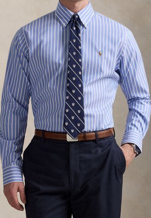 Homme portant une chemise rayée bleu et blanc avec boutons, une cravate bleu marine à motifs, une ceinture marron, un pantalon foncé et une montre-bracelet.