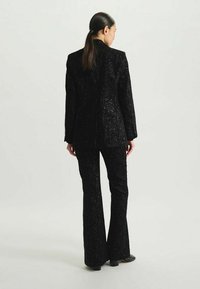 Costume noir pailleté comprenant un blazer ajusté et un pantalon évasé, avec un motif floral texturé et un design structuré.