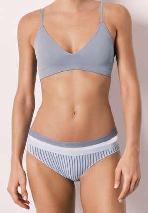 Femme portant un bralette côtelé bleu clair et une culotte rayée assortie avec des ceintures de marque, debout devant un fond uni.