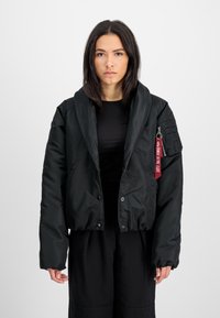 Svart cropped jakke med en jevn, glanset tekstur. Har stor krage, trykknappslukkinger og en glidelåslomme med et rødt nøkkelmerke.