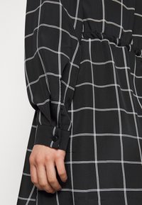 Černé kostkované šaty s bílou mřížkou, se staženým pasem, knoflíkovými manžetami a hladkým textilním povrchem.