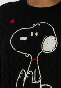 Un pull en tricot noir avec un chien cartoon brodé, des détails blancs et un cœur rouge accentué sur la gauche.