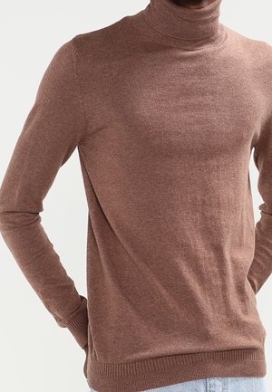 Mand iført en brun rullekravesweater med lange ærmer, stående med hænderne bag ryggen mod en ensfarvet baggrund.