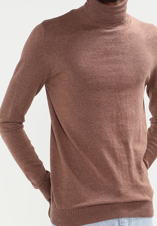 Homme portant un pull à col roulé marron à manches longues, debout les mains derrière le dos devant un fond uni.