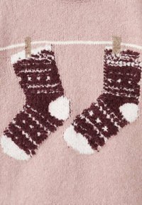 Pleteni pulover z dvema bakrenima teksturiranima nogavicama z belimi detajli, obešena na bež vrvici proti svetlo rožnati ozadju.